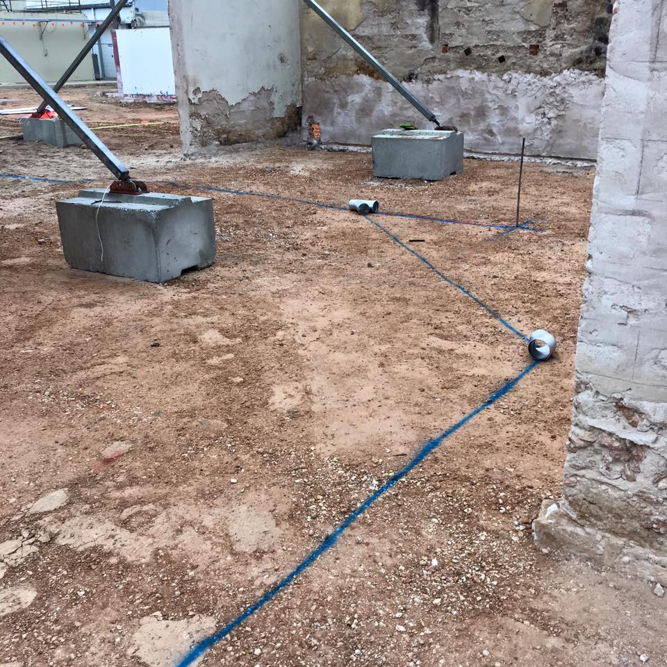 The image depicts a construction site where concrete bases are installed, likely to support new plumbing systems or structural elements. Steel supports, or struts, are visible, suggesting that this site may be undergoing significant renovations or new installations. The ground is uneven, covered with dirt and gravel, indicating ongoing site preparation. Blue lines on the ground appear to mark areas for plumbing or other utilities, likely indicating where pipes will be laid or other construction elements will be installed. This environment is relevant for Riverland Plumbing & Gas, as they might be involved in installing or connecting water and gas lines in this newly developed area. Such foundational work is crucial for ensuring the integrity and functionality of plumbing systems in both residential and commercial settings.