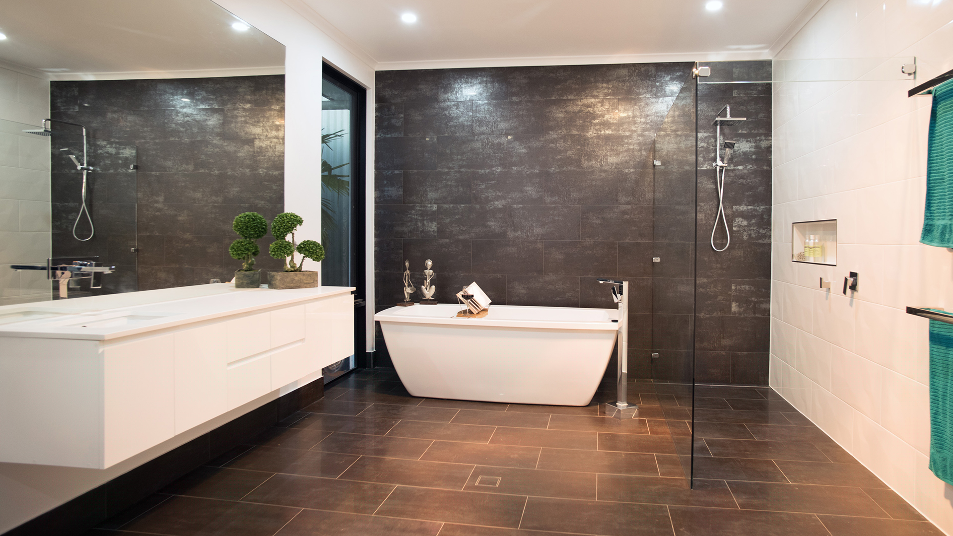 The image showcases a modern and stylish bathroom featuring a freestanding bathtub and a glass-enclosed shower. The walls are a combination of sleek dark tiles and bright white tiles, creating a contemporary aesthetic that is both sophisticated and inviting. Natural light pours in from a window, illuminating the room and highlighting the fixtures. A double sink vanity with a clean, minimalist design is situated on one side, while lush green plants add a touch of nature and freshness to the space. This setting exemplifies the high-quality plumbing work that Riverland Plumbing & Gas could provide, demonstrating their expertise in creating elegant and functional bathroom spaces for their clients.