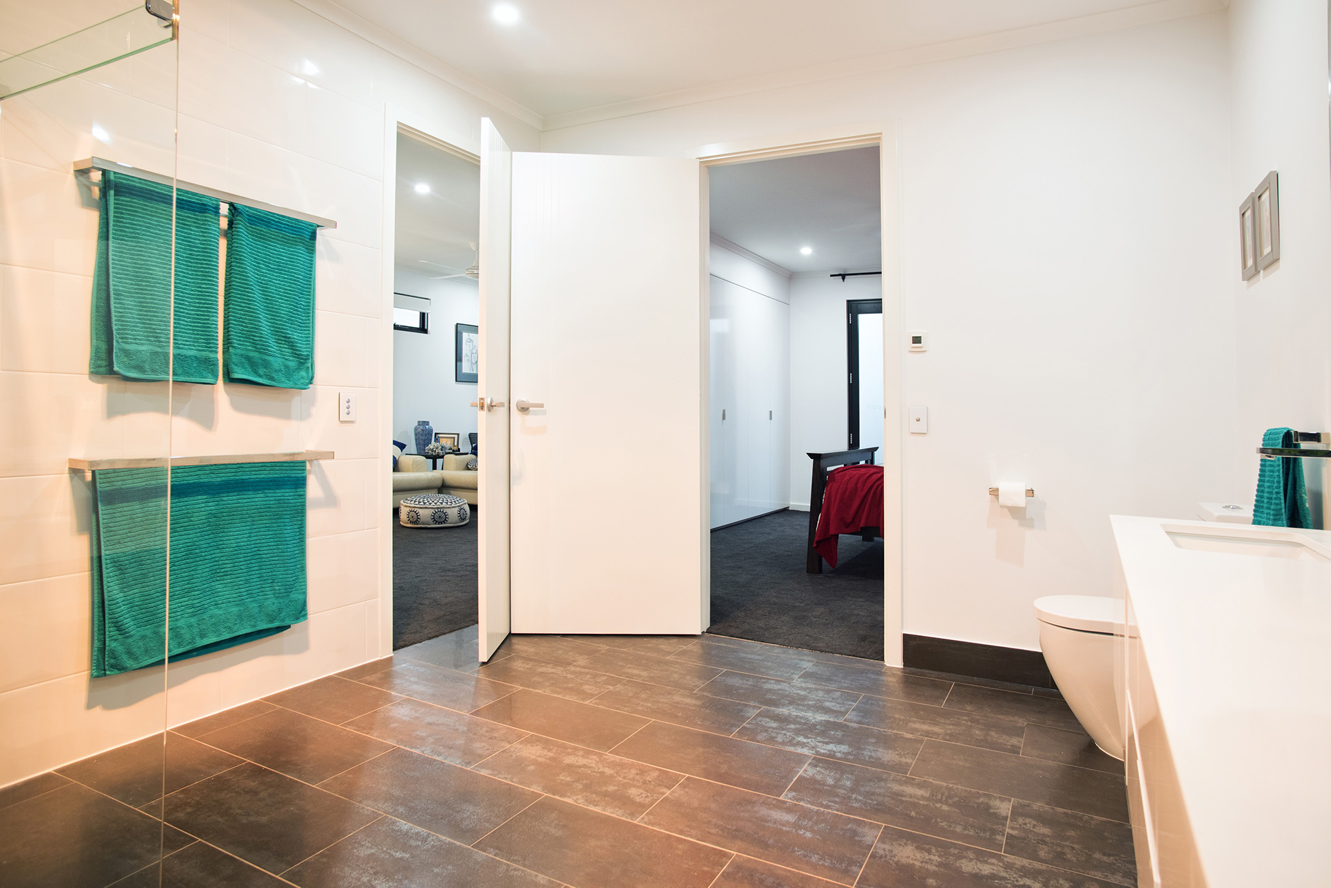 The image depicts a modern bathroom that is well-lit and features contemporary design elements. The space has glossy white tiles on the walls and a tiled floor in a darker shade, creating a stylish contrast. Two vibrant teal towels are neatly hung on chrome hooks on the wall, adding a pop of colour to the otherwise neutral palette. In the background, a door is partially open, revealing a glimpse of another room, while the bathroom has a clean and uncluttered look, indicating good maintenance. A toilet is positioned discreetly to the side, ensuring optimal use of space. This aesthetic suggests that the plumbing and fixtures are new and well installed, showcasing the quality of work that Riverland Plumbing & Gas provides. The focus on cleanliness and modern fixtures reflects the high standards of plumbing repairs and installations they offer in the Australian market.