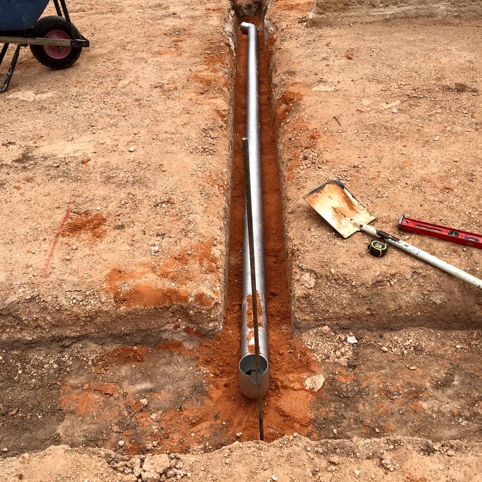 The image depicts a construction site with a trench excavated into the ground. In the centre, a long, shiny silver pipe is laid in the trench, suggesting that plumbing work is in progress. The sandy soil around the trench indicates that the area may be in the early stages of site preparation for construction or renovation. To the side, a wheelbarrow and tools including a shovel and a wrench are visible, emphasising that this site is actively being worked on. This scenario aligns perfectly with Riverland Plumbing & Gas's services, indicating that they might be installing or repairing underground plumbing systems as part of their expertise in providing efficient plumbing solutions across Australia.
