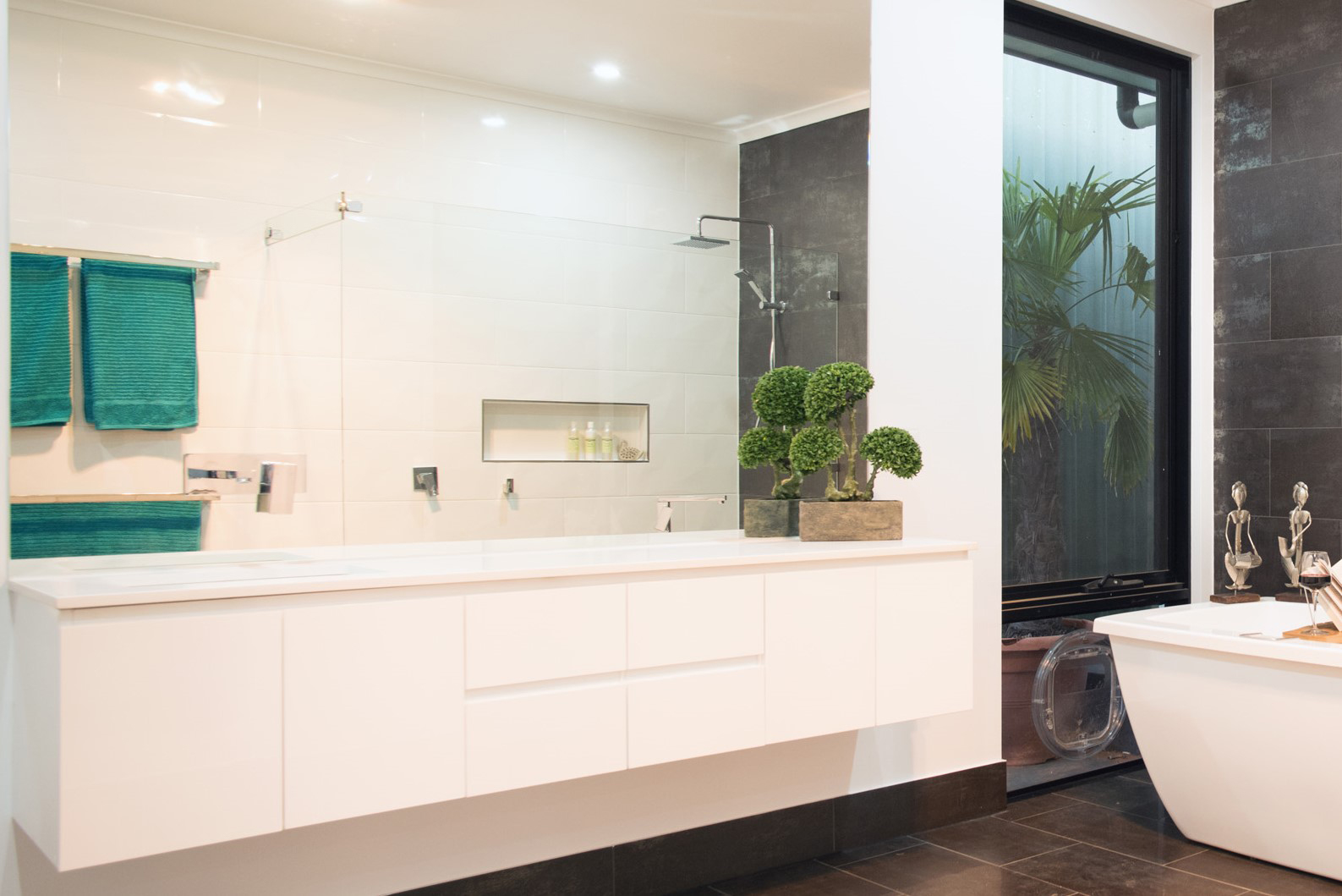 The image showcases a modern bathroom featuring a sleek design with contemporary elements highlighting the skills of Riverland Plumbing & Gas. The bathroom includes a large, white vanity with multiple drawers for storage, topped with a minimalist basin. To the left, there is a glass shower enclosure with modern fixtures, ensuring a premium showering experience. The walls are adorned with elegant white tiles, and an eye-catching green accent is provided by the rolled towels hanging on a stylish rack. Additionally, a potted plant adds a touch of nature to the space, contributing to a fresh and inviting atmosphere. The overall aesthetics reflect high-quality craftsmanship and attention to detail, offering potential customers a glimpse of what Riverland Plumbing & Gas can achieve in bathroom renovations or plumbing installations.