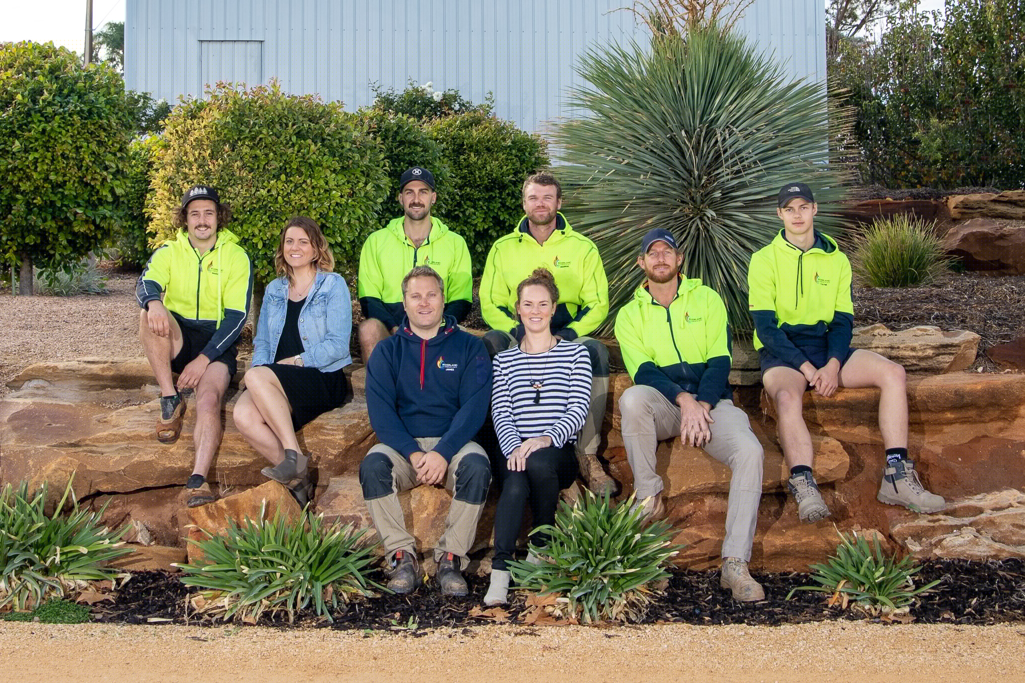 The image features a group of individuals associated with Riverland Plumbing & Gas, positioned outdoors on a natural stone lush setting. The group consists of eight people, appearing friendly and approachable. Seven of the team members are dressed in bright yellow high-visibility work shirts, indicative of their plumbing and gas services, and they are seated or standing together on a large rock formation. Among them is a woman wearing a casual outfit, standing slightly apart but still integrated into the group, which conveys a sense of teamwork and community. In the background, there are various shrubs and plants, enhancing the outdoor atmosphere. The overall composition reflects a professional yet friendly image of the plumbing team, showcasing their unity and readiness to serve the community in Australia. This setting symbolizes reliability and professionalism in the plumbing industry, aligning with the values of Riverland Plumbing & Gas.