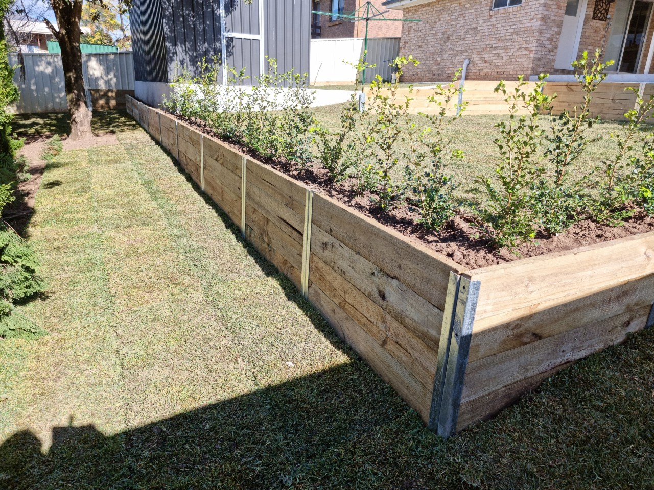 The image showcases a well-maintained garden featuring a wooden retaining wall. The wall is constructed with timber planks and is reinforced with metal supports. Behind the wall, there are neatly planted shrubs, and the surrounding area features freshly mowed grass, creating a tidy and appealing outdoor space.