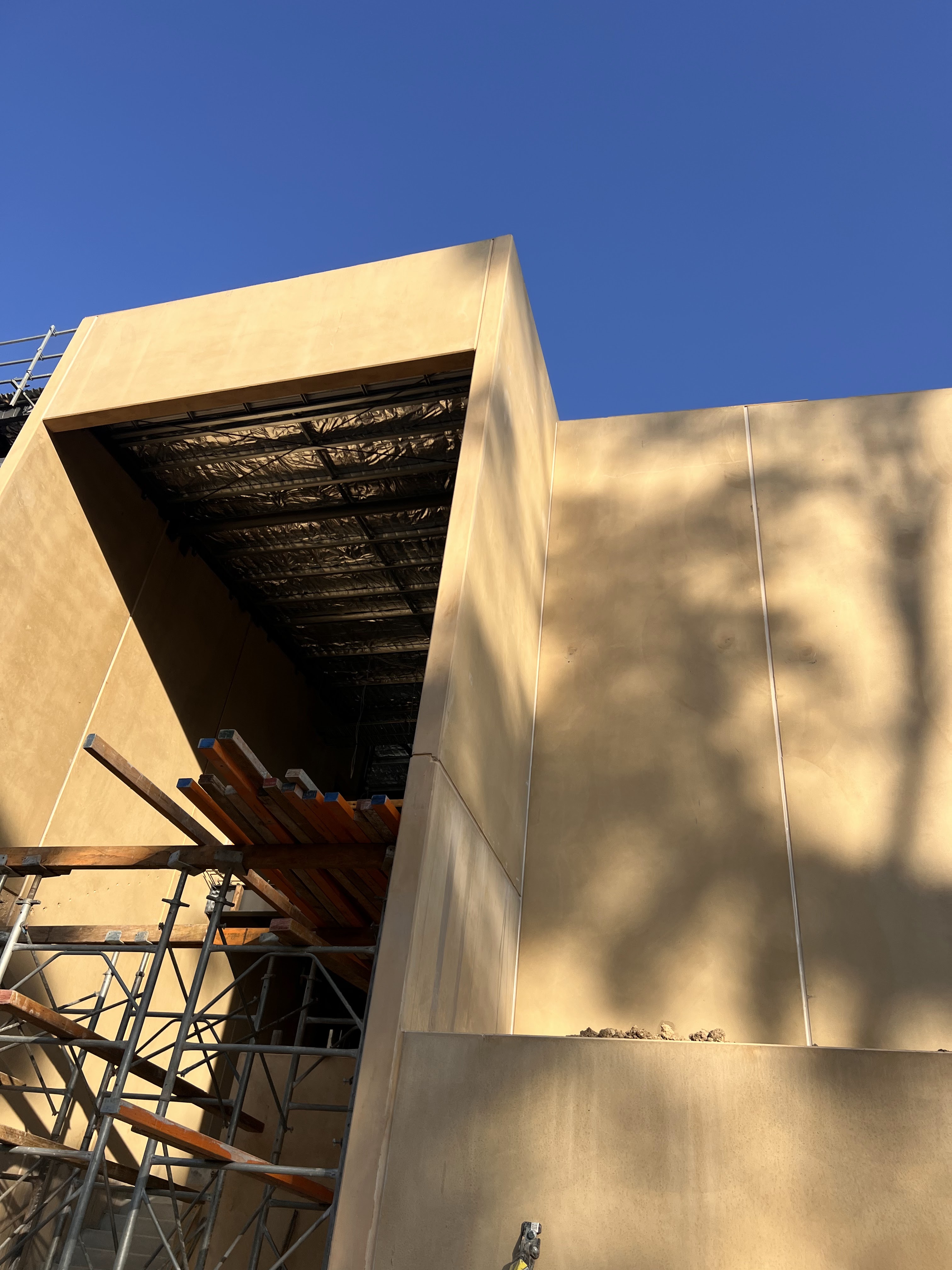 The image showcases a partially constructed building featuring a distinct geometric design. The surface of the main wall exhibits a smooth, tan-coloured finish under a clear blue sky. A sizeable opening within the structure hints at modern architectural elements, suggesting the addition of glass or another material in the future. Surrounding the building, scaffolding is visible, indicating ongoing construction work. The use of construction tools and materials in view reinforces the active building site atmosphere.