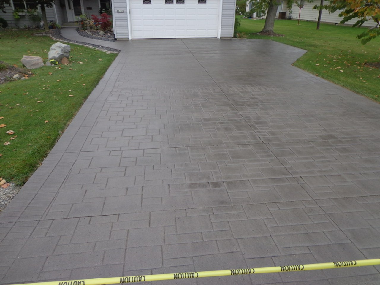 The image features a newly paved driveway with a smooth, grey surface, showcasing a brick-like pattern. Surrounding the driveway is a well-maintained lawn with green grass and some rocks, framed by trees.
