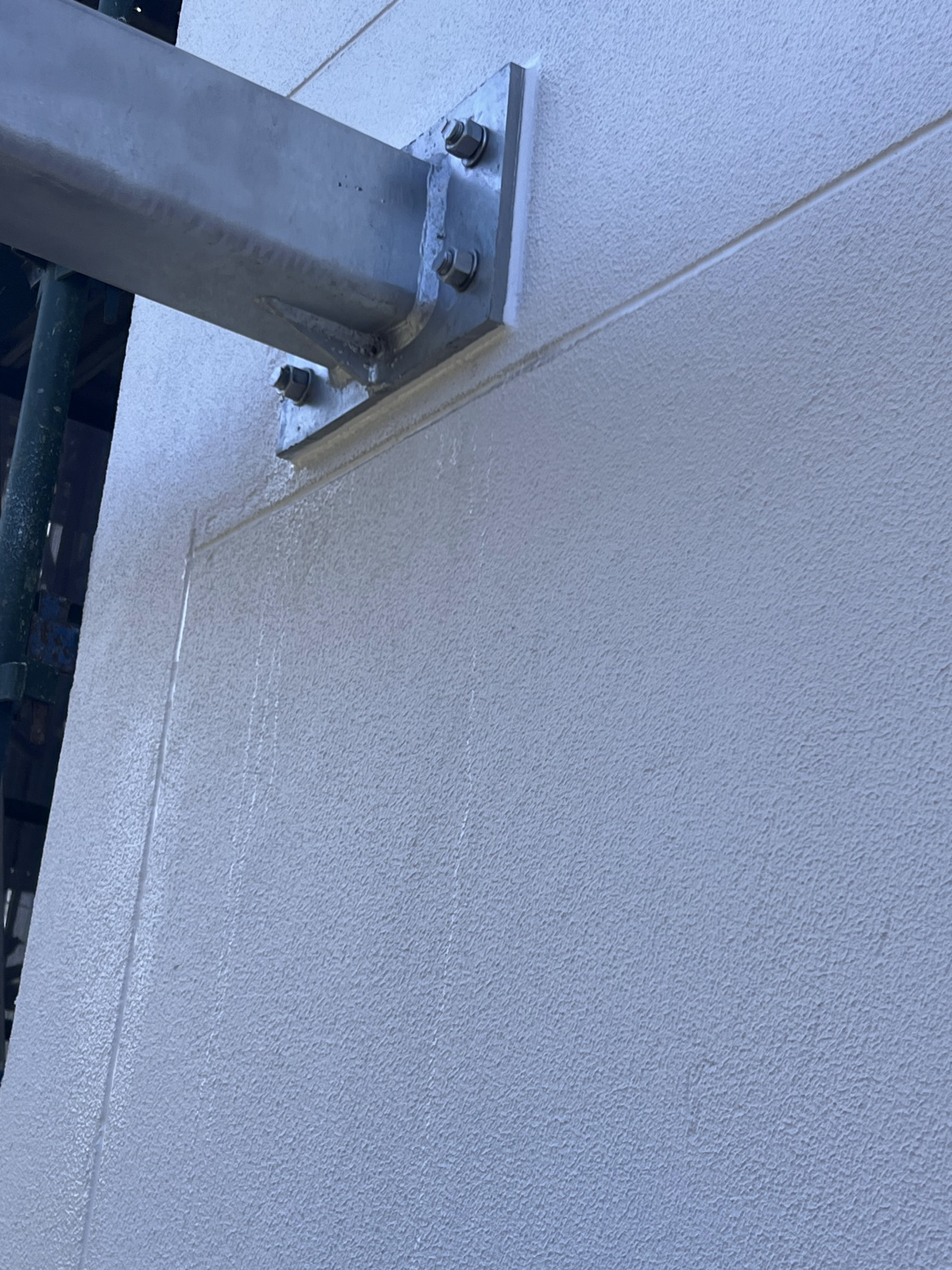 The image presents a close-up view of a textured wall section, which appears to be part of a construction or renovation project. A structural beam or pipe is visibly attached to the wall, with visible mounting brackets and bolts. There is some water streaking down the wall, indicating potential weather exposure or construction activity. The wall's surface appears to be smooth but shows signs of moisture, suggesting ongoing work or a recent installation. This scene is indicative of the types of services offered by Skill CPG Construction Pty Ltd, focusing on structural integrity and finishing details in construction projects.