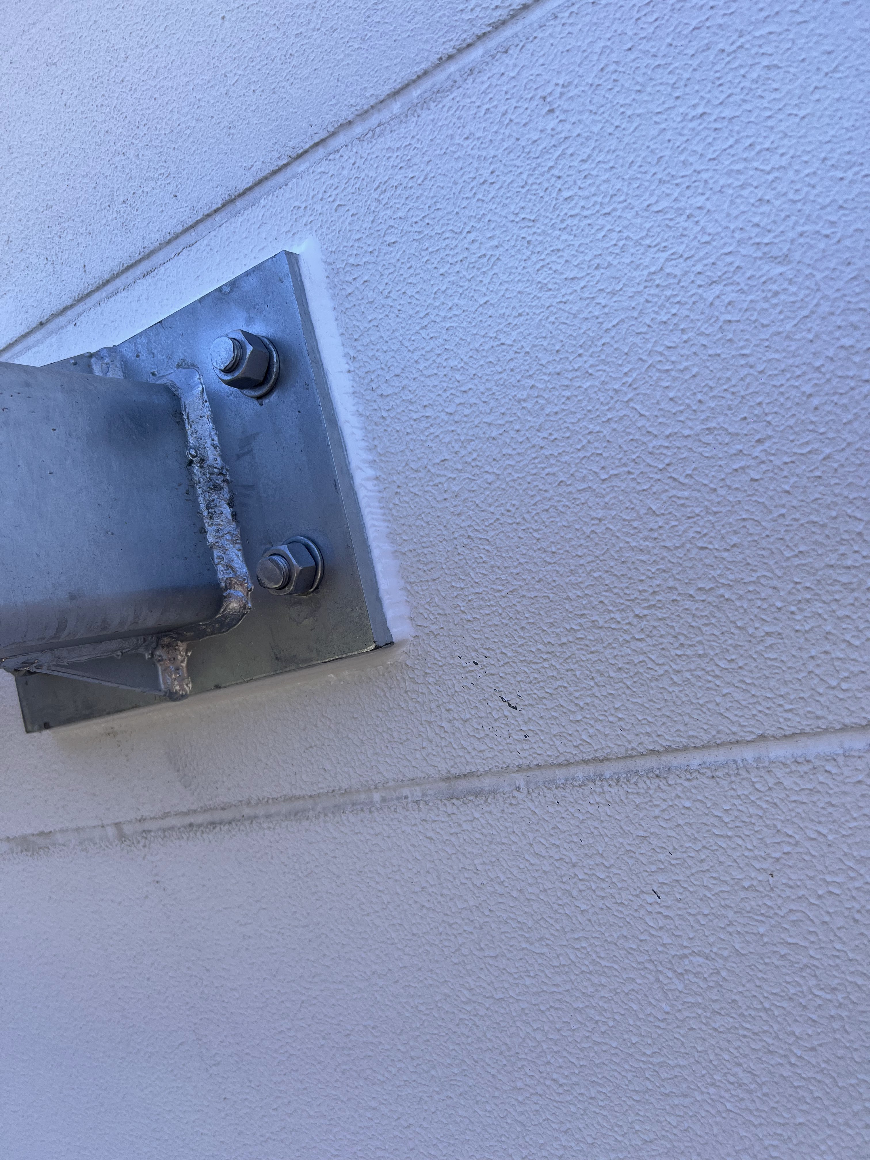 The image shows a close-up view of a metal bracket attached to a textured white wall. The bracket appears to be used for structural support, fixed with bolts. The clean lines and metal surface suggest a professional and durable installation, common in construction. The surrounding wall is smooth but exhibits some minor markings, possibly from previous installations or a standard wear-and-tear. This element showcases attention to detail in building practices, emphasising the importance of robust structures in construction projects.