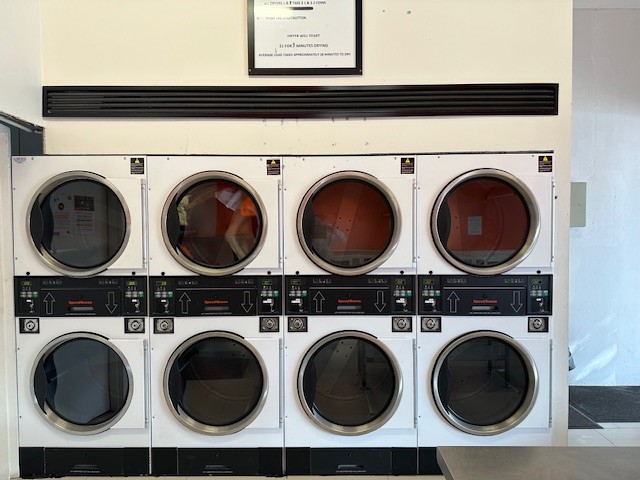The image shows a row of six industrial drying machines, all featuring large circular glass doors and clearly marked control panels. The machines are clean and modern, reflecting a professional dry cleaning and laundromat environment. The walls behind the machines are plain, suggesting a tidy and well-maintained facility, ideal for customers looking for efficient dry cleaning services. Overall, the setup indicates a focus on quality service in the laundry process, catering to various clientele in Attadale and surrounding areas.