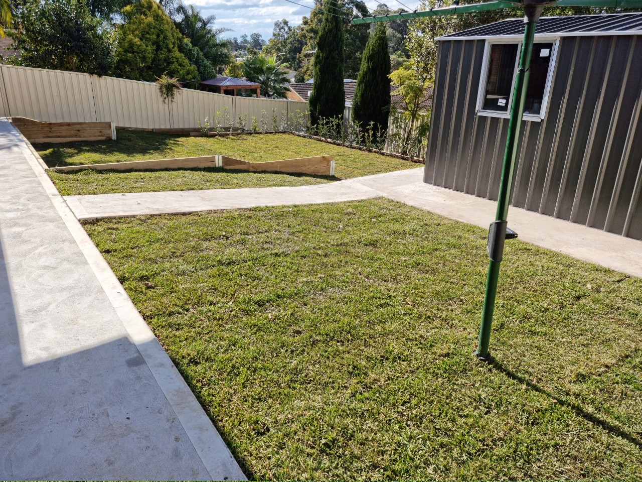 The image showcases a well-maintained garden area with freshly laid grass, bordered by concrete pathways. In the background, there is a modern shed with a metallic finish, surrounded by trees and neatly arranged landscaping. The area looks tidy, with a clear delineation between the grass and the paved surface.