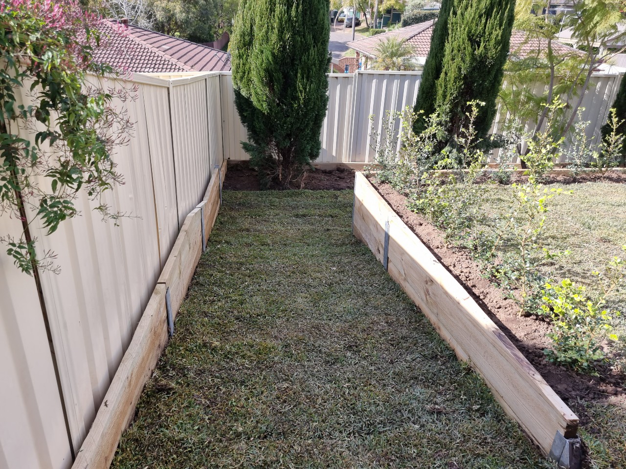 The image showcases a well-maintained garden pathway bordered by wooden retaining walls. The path is lush with green grass, leading through a tidy landscape featuring neatly trimmed shrubs and small plants on either side. The overall atmosphere is serene and organised, with clear boundaries created by the wooden structures.