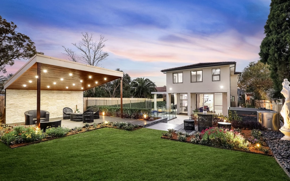 The image features a beautifully landscaped garden with a well-maintained green lawn. In the foreground, there is a modern outdoor seating area with stylish chairs under a wooden pergola adorned with lights. The background showcases a contemporary two-storey house with large windows. Flower beds with colourful plants and shrubs line the garden, adding vibrant touches. There are also trees and a well-defined pathway leading to the entrance of the home, creating an inviting atmosphere.