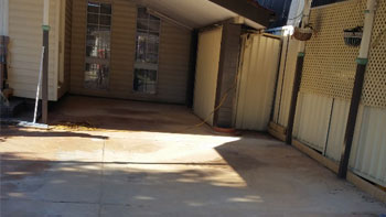 The image shows a spacious area with a concrete surface, partially shaded by a slanted roof. There are wooden doors visible on one side, and a fence with a pale yellow colour runs along the perimeter. The space appears clean and ready for potential landscaping or outdoor use.