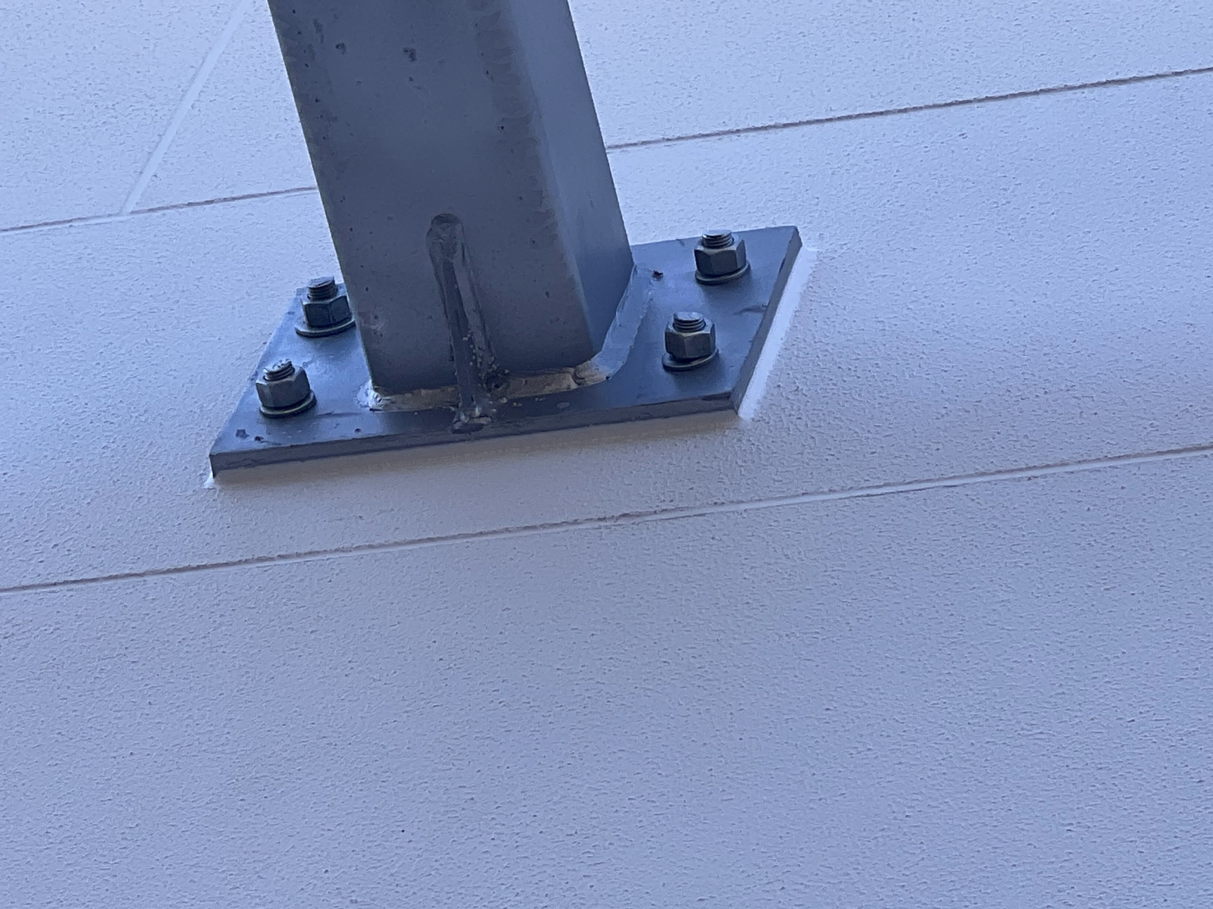 The image depicts a steel column baseplate secured to a wall, featuring several bolts fastening it in place. The baseplate appears to be integral to a construction project, likely supporting structural elements above. The clean finish and modern materials suggest it is part of a contemporary building project, suitable for commercial or industrial use. Proper installation and bolting indicate adherence to safety and engineering standards, essential in construction.