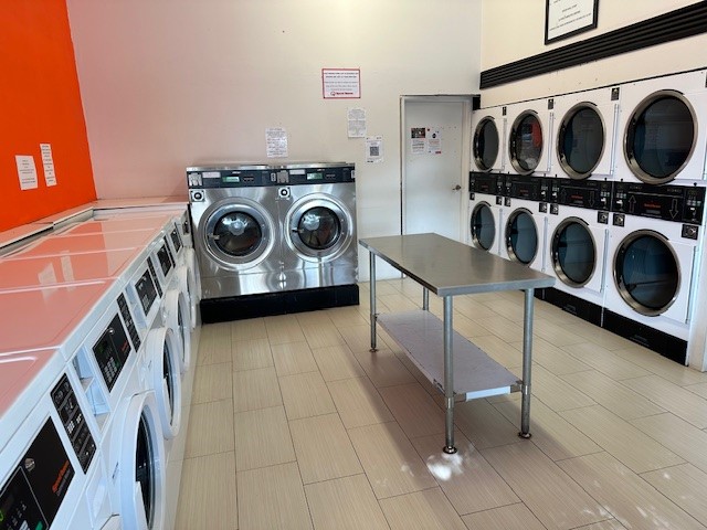 The image depicts the interior of a laundromat. On one side, there are several washing machines lined up, featuring modern designs and digital displays. The machines have a clean, shiny appearance, suggesting they are well-maintained. Adjacent to the washing machines, there is a folding table in the centre of the room, designed for customers to conveniently fold their laundry after washing. On the opposite wall, there are high-efficiency dryers, similarly modern in design, installed in a row. The walls are painted a vibrant orange, contrasting with the white and stainless steel of the appliances. There are notices and instructions visible, likely providing information on the washing and drying services available, indicating a customer-friendly environment.