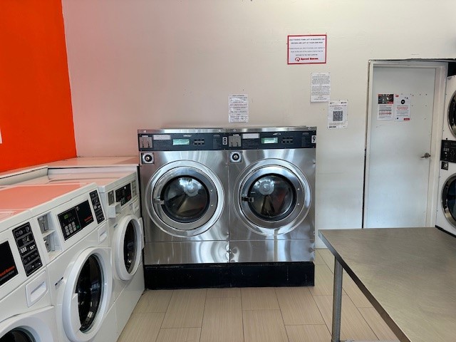 The image shows the interior of a laundromat service with a focus on two large, shiny washing machines that are placed side by side. These machines are equipped with digital displays for selecting wash cycles. To the left, there are multiple smaller washing machines arranged in a row, all having similar digital interfaces. The overall setting is bright, enhanced by an orange wall, suggesting a welcoming atmosphere for customers. There’s also a stainless steel table in the foreground, likely used for folding clothes or providing service assistance. The cleanliness and modernity of the machines indicate that this laundromat takes pride in its equipment, contributing to an efficient dry cleaning experience for customers in the nearby areas.