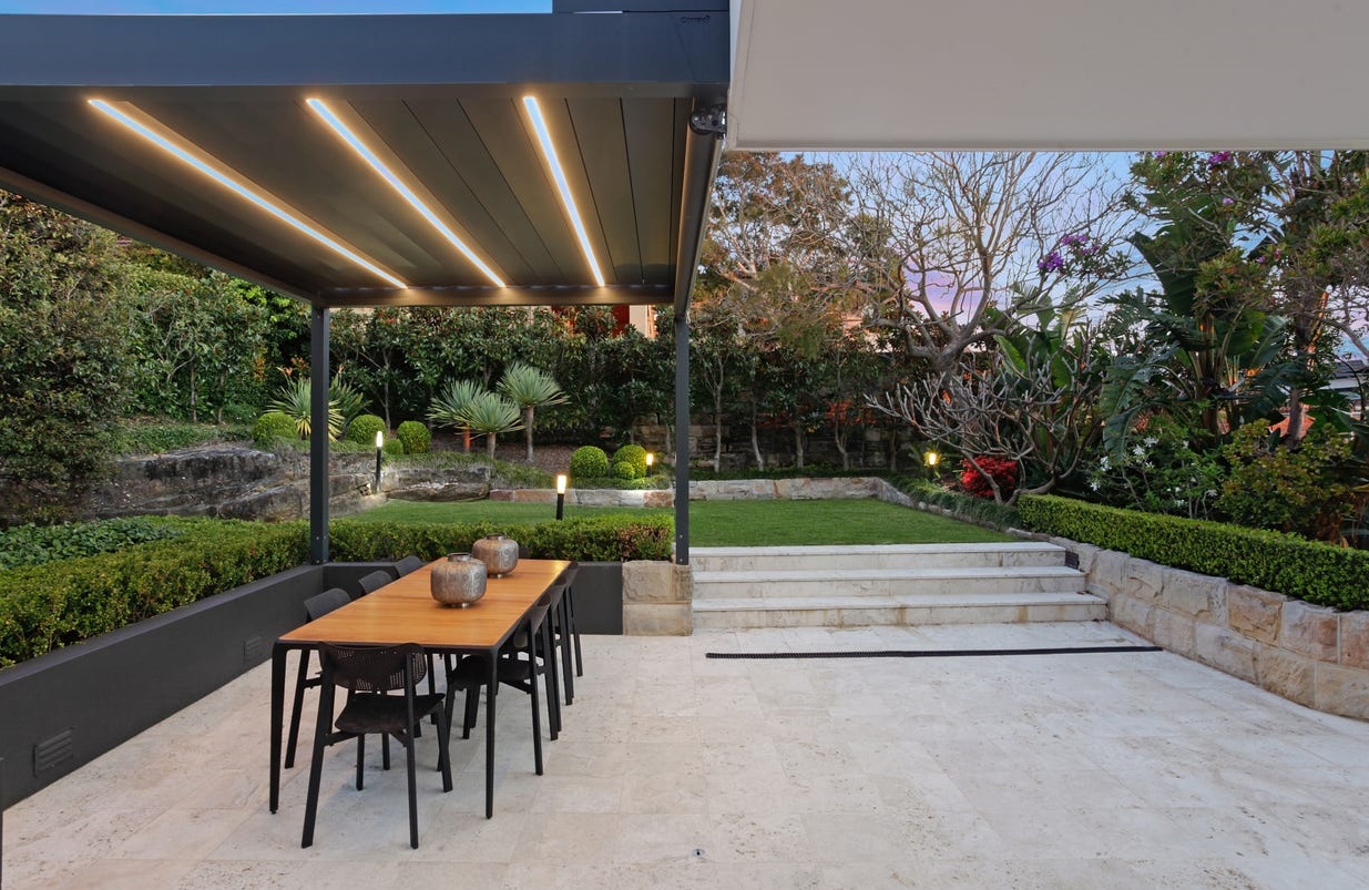 The image showcases a beautifully designed outdoor space featuring a modern dining area with a wooden table and black chairs, situated under a sleek pergola with LED lighting. Surrounding the area are well-maintained green hedges and various ornamental plants, creating a serene garden atmosphere. Steps lead to a lush lawn that is bordered by vibrant landscaping, including flowering plants and sculptural foliage, all set against a tranquil evening sky.