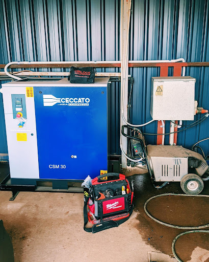 The image features a blue industrial compressor, branded 'Ceccato', accompanied by a red tool bag, likely containing electrical tools. The compressor is located in a workshop or industrial setting, with visible pipes along the walls indicating an operational workspace. The image suggests a focus on electrical or pneumatic tools commonly used by electricians, highlighting the equipment relevant to the profession.