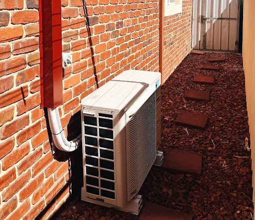 The image features an external air conditioning unit positioned against a brick wall. It is located in a narrow pathway surrounded by reddish gravel. There appears to be a drain pipe connecting to the unit, and the area is well-maintained with stepping stones leading towards a gated entry at the end of the pathway. The utilisation of bricks suggests a rustic aesthetic, suitable for residential or commercial properties. This type of installation is often relevant for electricians involved in HVAC systems or energy-efficient upgrades.