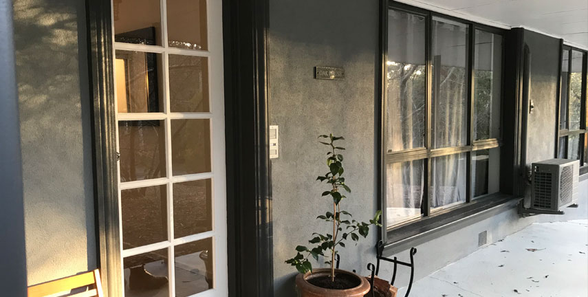 The image depicts an entrance to a house featuring a set of French doors framed by a grey wall. The porch has a small potted plant and a chair, with large windows allowing natural light to illuminate the space. The exterior is modern and inviting.