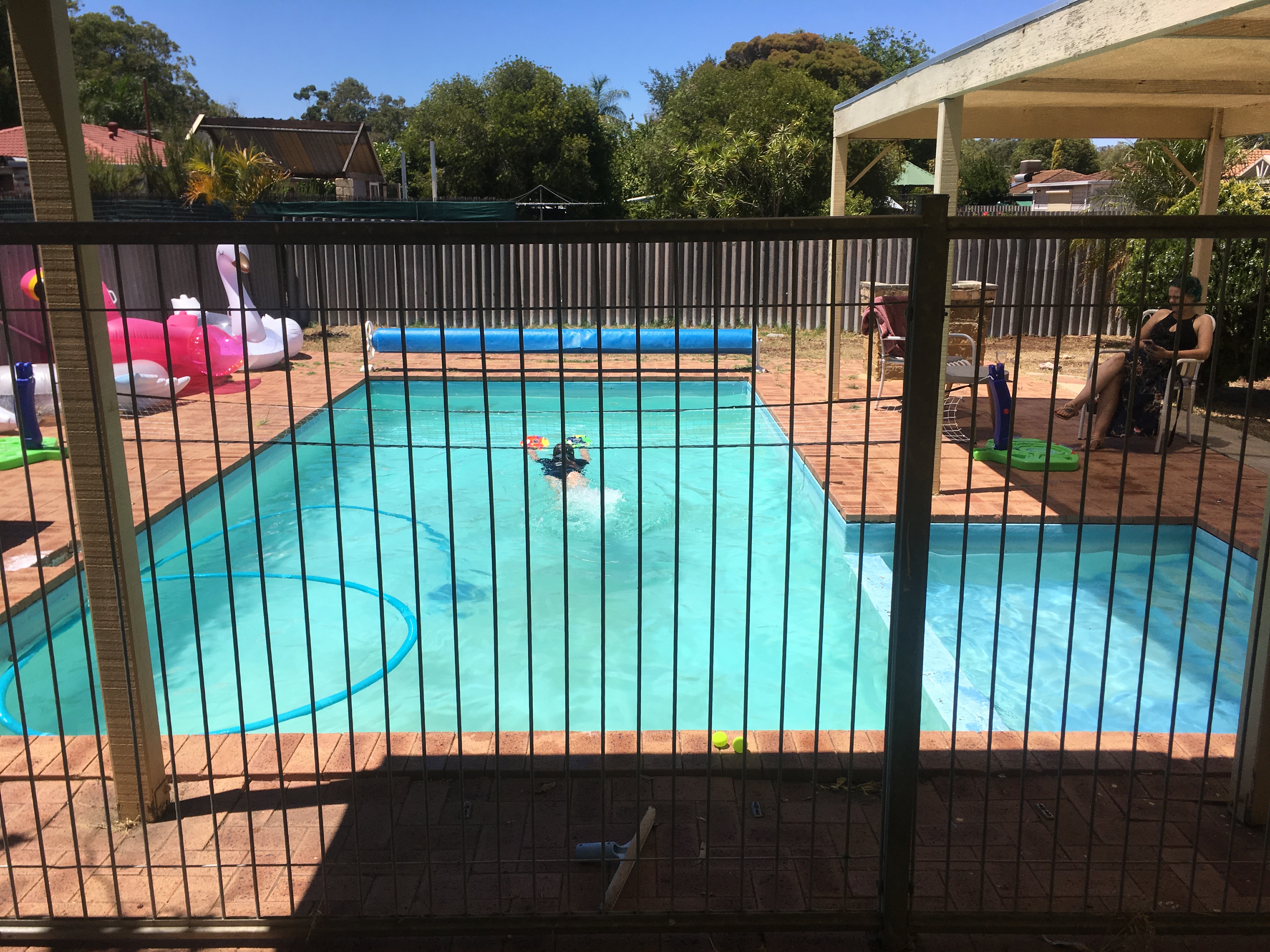 The image shows a residential swimming pool with a turquoise surface, surrounded by a brick-paved area. The pool features items such as inflatables, including a large pink flamingo, and a floating lounge. In the centre, a child can be seen playing in the water, holding a colourful toy. The pool is enclosed by a safety fence, and in the background, there are several trees and residential buildings. To the right, two adults are seated on chairs, enjoying the sun, adding a relaxed atmosphere to the scene.