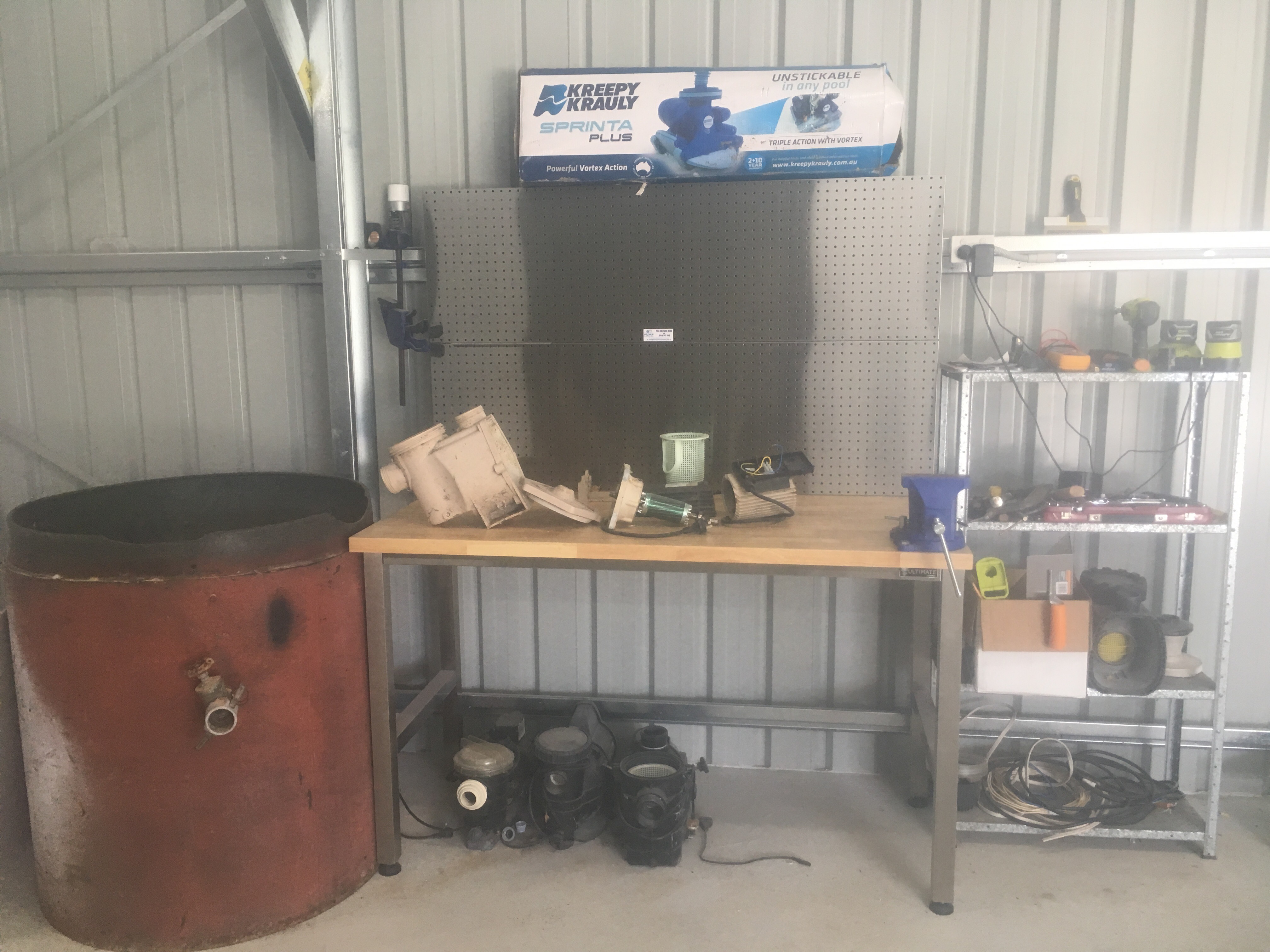 The image shows a workspace set up for pool repairs. There is a large red tank on the left, possibly for storing equipment or chemicals. A sturdy stainless steel table occupies the middle, laden with various pool repair tools and equipment, including pumps and other pool accessories. Up on the wall, there's a pegboard that appears to have additional tools and organisational items attached for easy access. In the background, a banner for "Kreepy Krauly" can be seen, indicating the focus on pool cleaning equipment. This setup illustrates an organised and functional environment for a swimming pool repair service.