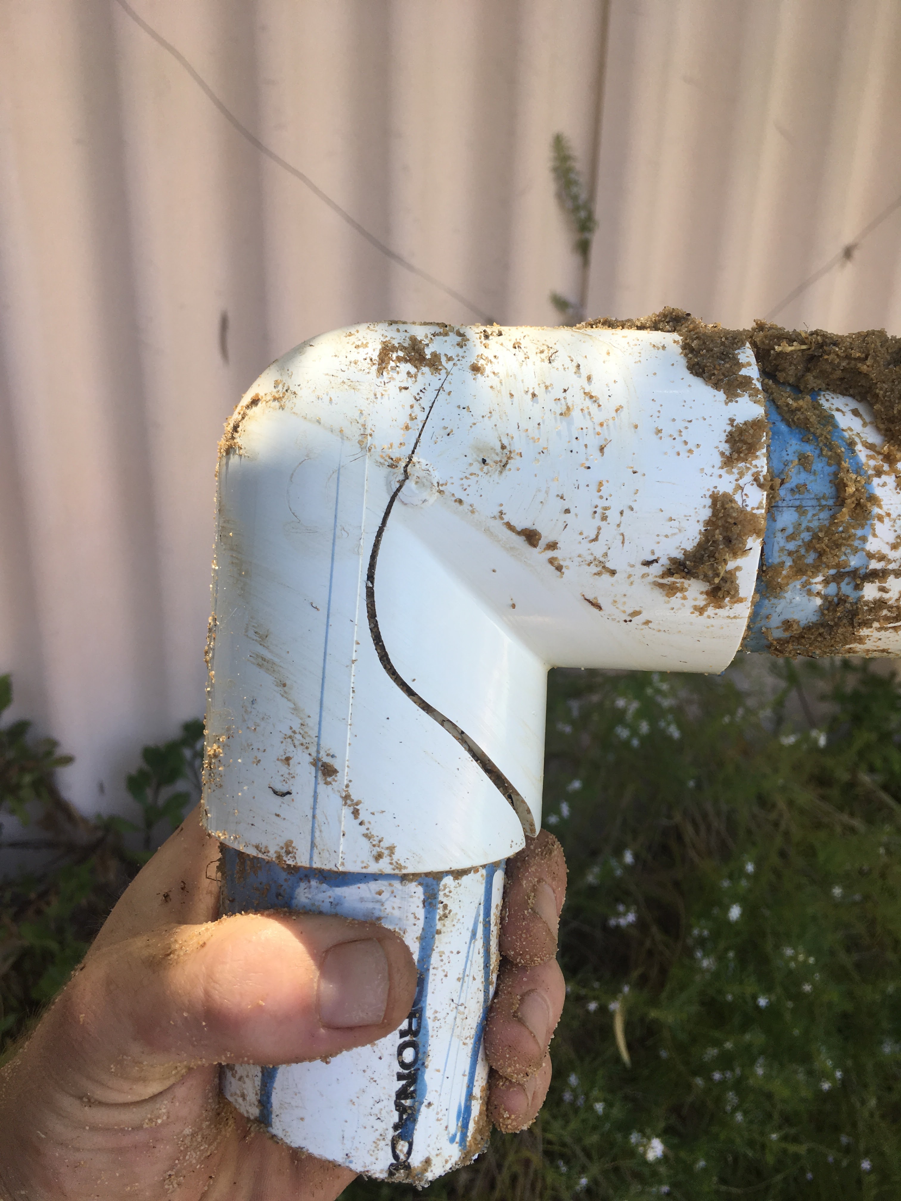 The image shows a close-up view of a white PVC pipe fitting with elbow joints, which are commonly used in swimming pool plumbing systems. The pipe is dirty and covered in sand, indicating recent excavation or maintenance work. It appears that the pipe is being inspected or prepared for repair, which relates to the services offered by Perth Pool Pump Repairs. The surrounding vegetation suggests this might be an outdoor area, perhaps near a swimming pool. Properly functioning plumbing is essential for maintaining a swimming pool, highlighting the importance of repair and maintenance services.