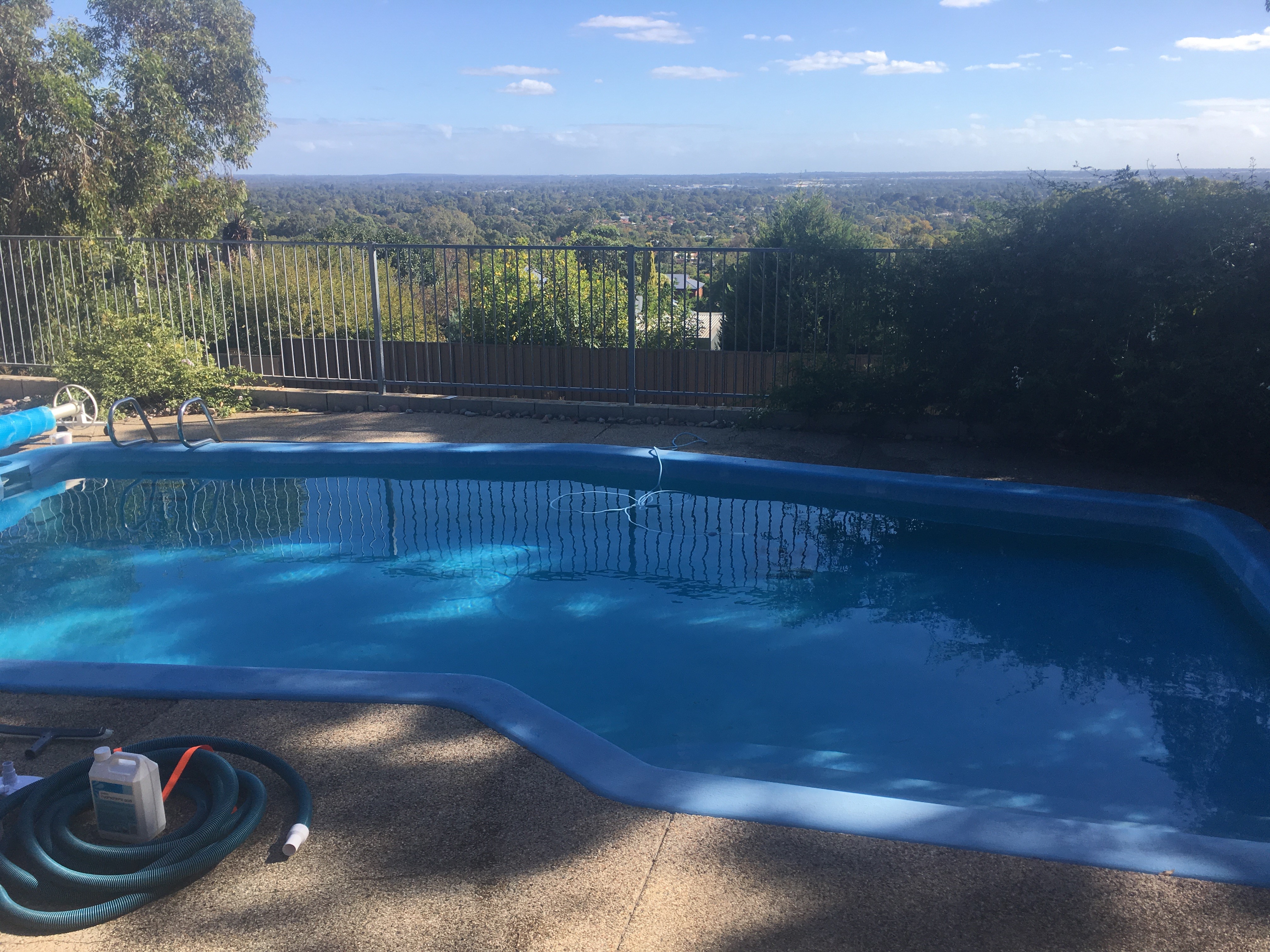 The image depicts a clear swimming pool, surrounded by a well-maintained area. The pool features a light blue interior, and the water is sparkling, indicating cleanliness and proper maintenance. In the background, greenery is visible, along with a stunning panoramic view of the landscape beyond the pool area. Sunlight shines brightly on the water, creating a serene ambiance.