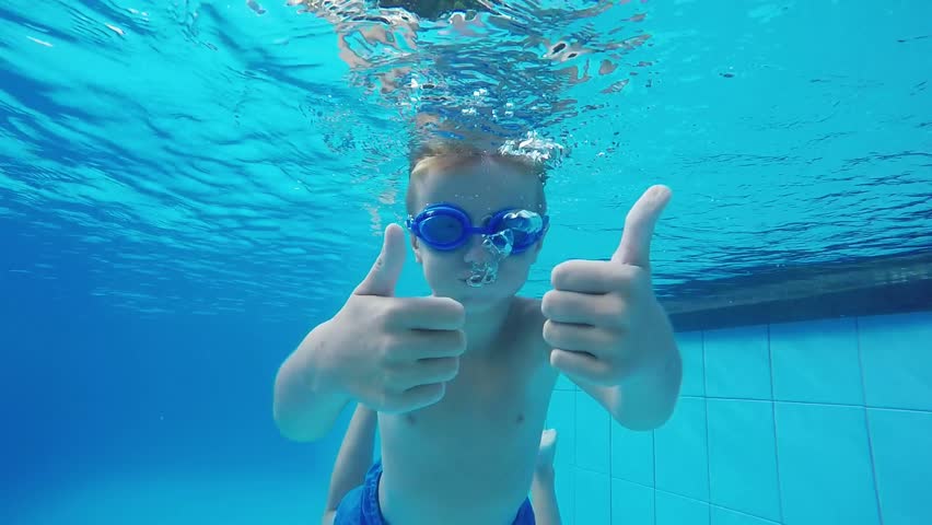 The image depicts a young child submerged in a swimming pool, giving two thumbs up while wearing blue swimming goggles. The water is clear and vibrant, casting a bright blue hue around the child, enhancing the lively atmosphere typical of a summer day at the pool.