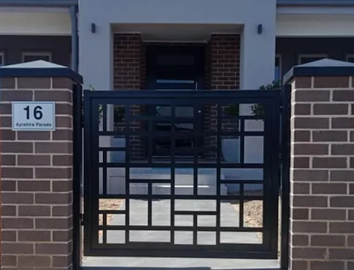 The image depicts a contemporary property entrance with a decorative metal gate set between two brick pillars. The gate features a geometric design, showcasing a modern aesthetic that aligns well with smart home solutions. The neatly paved pathway leads up to the main entrance of the house, which is visibly well-maintained, reflecting a stylish and secure home environment. The house's exterior comprises a mix of brick and render, indicating a modern architectural style. This setup suggests a focus on enhancing security and aesthetics, pertinent for a home automation company like Southern Gate Automation.