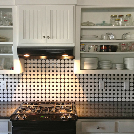 The image showcases a modern kitchen featuring a sleek black stove beneath a stylish range hood. The backsplash is adorned with circular black and white tiles, creating a striking visual effect. Above the stove, there are white cabinetry with glass doors displaying various glassware and dishes. The countertop is dark and polished, providing contrast to the lighter cabinetry.