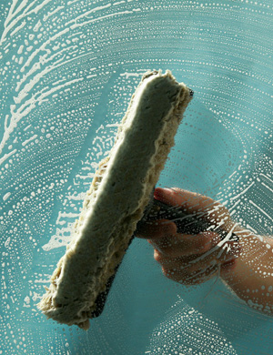 The image features a close-up of a hand holding a squeegee against a glass surface that is being cleaned. The surface has soap suds and streaks, indicating that the cleaning process is underway. The background is a soft blue tone, which enhances the impression of a fresh and clean environment. This scene aligns perfectly with House Cleaning Cronulla's operations, showcasing the professionalism and attention to detail that the business provides in their commercial cleaning services. The use of a squeegee suggests an efficient approach to window cleaning, highlighting the thoroughness of their services.