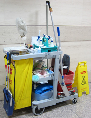 The image displays a cleaning cart that is commonly used in commercial cleaning services. The cart is equipped with various cleaning supplies and tools essential for maintaining cleanliness in commercial spaces. It features compartments filled with bottles of cleaning solutions, sponges, cloths, and mops. A yellow caution sign is visible, warning of wet floors, indicating that cleaning is in progress. This is typical of the House Cleaning Cronulla business, as they offer professional cleaning services in locations such as Caringbah, Yowie Bay, and Woolooware. The cart's arrangement reflects an efficient approach to commercial cleaning, facilitating quick access to necessary tools and enhancing productivity as their team services various establishments in the area.