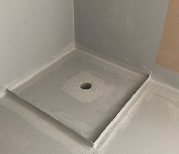 The image shows a freshly completed waterproofing installation in a wet area, likely intended for a shower base or a similar application. The surface appears smooth and finished, with a drain hole centred in the middle of a rectangular base that has been prepped for waterproofing. The walls surrounding the area are also treated, indicating readiness for tiles or other surface finishes. This stage of waterproofing is crucial to prevent leaks and water damage in residential or commercial spaces.
