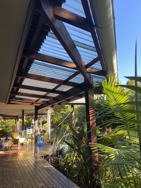 A well-crafted timber pergola with a transparent corrugated roof attached to a house, providing a shaded outdoor area. The wooden deck beneath is surrounded by lush greenery, highlighting expert carpentry work in an outdoor living space typical of Brunswick Heads and nearby areas.
