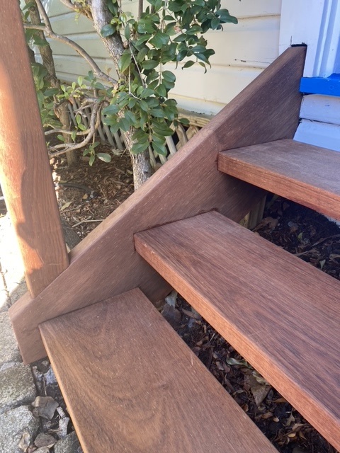 The image shows a close-up view of a wooden staircase, likely custom-built, featuring smooth, polished timber steps and a sturdy wooden stringer supporting the structure. The craftsmanship highlights precise joinery and finishing, set outdoors next to a white painted wall with a blue trim detail. Surrounding foliage and natural ground elements suggest the stairs are part of an outdoor area in a residential setting in Brunswick Heads, NSW.