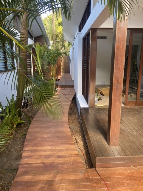 The image shows a beautifully crafted wooden walkway and deck area adjacent to a modern white building. The walkway is made from smoothly finished timber planks with a natural warm tone, winding alongside lush green plants and leading to a wooden deck supported by sturdy timber posts. The deck appears to be part of an outdoor living space with large glass doors providing access to the interior. The carpentry work is precise, highlighting quality timber finishing and an elegant design suited for residential properties in the Brunswick Heads area.
