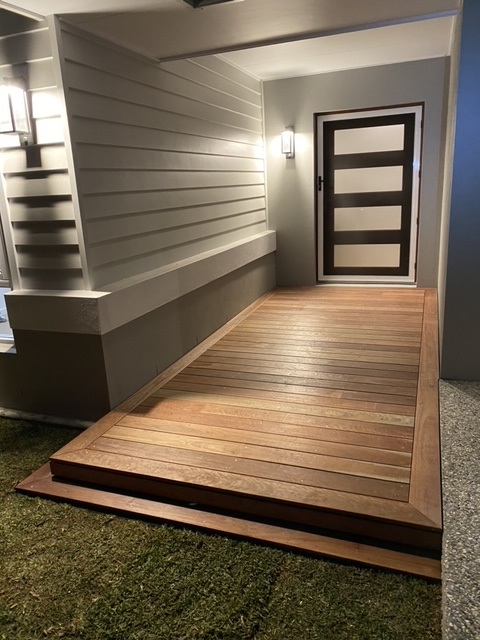 The image showcases a beautifully crafted wooden ramp leading to a modern front door, illuminated by exterior wall lights. The ramp is constructed with smooth, polished timber planks neatly bordered by a darker wood trim, blending seamlessly with the house's contemporary design. The surrounding area includes a patch of grass and a paved driveway, highlighting the ramp as a functional and aesthetic addition to the entrance, expertly demonstrating carpentry skills suitable for homes in Brunswick Heads and surrounding areas.