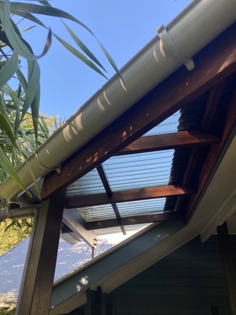 The image showcases a well-crafted wooden pergola structure with a transparent corrugated plastic roof panel, attached to a house exterior. The wood beams display a rich, polished finish, highlighting skilled carpentry work. The roof gutter is neatly installed above the wooden framework. Surrounding foliage and clear blue sky complement the outdoor setting, reflecting a blend of functional and aesthetic carpentry.