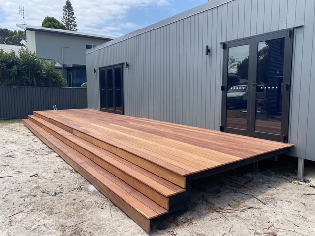 The image shows a newly constructed, elegant wooden deck with multiple steps leading up to a modern grey-clad building. The deck is made from polished timber, showcasing fine carpentry skills, suitable for outdoor residential projects. The clean and smooth finish highlights expert craftsmanship likely offered by Carpentry Byron Bay, serving the Brunswick Heads area and surroundings.