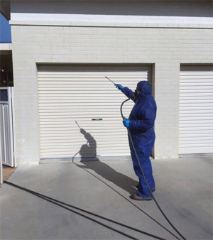 A pest control technician from Bolton Pest Control is dressed in full protective gear, including a blue coverall, gloves, and a face mask. He is using a spraying device to treat the exterior of a white residential garage door, presumably for pests. The location appears to be a residential area in Leeton, NSW, where the company operates. The shadow on the garage door highlights the technician's action, emphasising the pest control process.