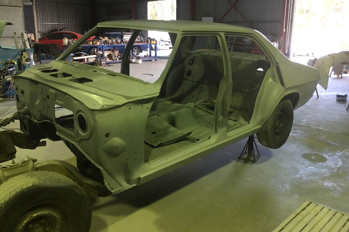 The image showcases a stripped-down car body, likely in the process of restoration or preparation for repainting. The body is mounted on stands, indicating it's being worked on in a workshop environment. Surrounding tools and equipment can be seen in the background, indicating a professional panel beating and painting setup. The overall condition of the car body appears to be in preparation for further refinements, highlighting the attention to detail typical in custom panel beating services.