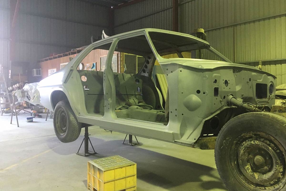 The image showcases a car shell in the midst of restoration, positioned on stands within a workshop. It appears to be stripped down, ready for bodywork. The environment suggests a dedicated panel beating and car painting facility, indicative of Rapley's Custom Pain & Panel's operations. Tools and equipment can be faintly seen in the background, highlighting the attention to detail and craftsmanship involved in the vehicle restoration process.