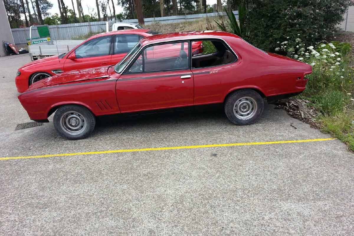 The image features a red vintage car parked in a lot within a car body shop setting. The vehicle is positioned next to a second car, which appears to be a modern sports model. The background shows greenery and foliage, indicating a well-maintained area. The car seems to require some restoration, typical for classic models needing refurbishment. This imagery could attract customers looking for panel beating and painting services for vintage or classic vehicles.