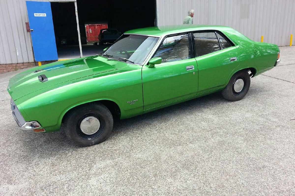 The image features a vintage green sedan parked on a concrete surface, likely outside a car body shop. The background suggests an industrial environment, with a blue door and another vehicle faintly visible in the distance. This vehicle represents the kind of cars that might be serviced or restored at Rapley's Custom Pain & Panel, highlighting their expertise in handling classic vehicles and custom paint jobs.