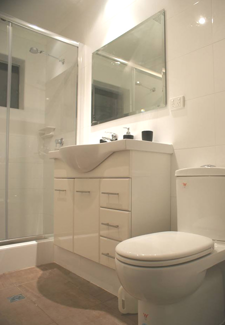 The image showcases a modern bathroom featuring clean, sleek design elements. It includes a glass shower enclosure, a white vanity unit with storage drawers, a matching white toilet, and a large mirror that reflects ambient lighting. The tiles are glossy and light-coloured, contributing to a spacious and airy feel. This contemporary setup illustrates a well-maintained plumbing installation, ideal for showcasing the quality work that Assurance Plumbing & Gas can provide in areas like Wanneroo and surrounding suburbs.