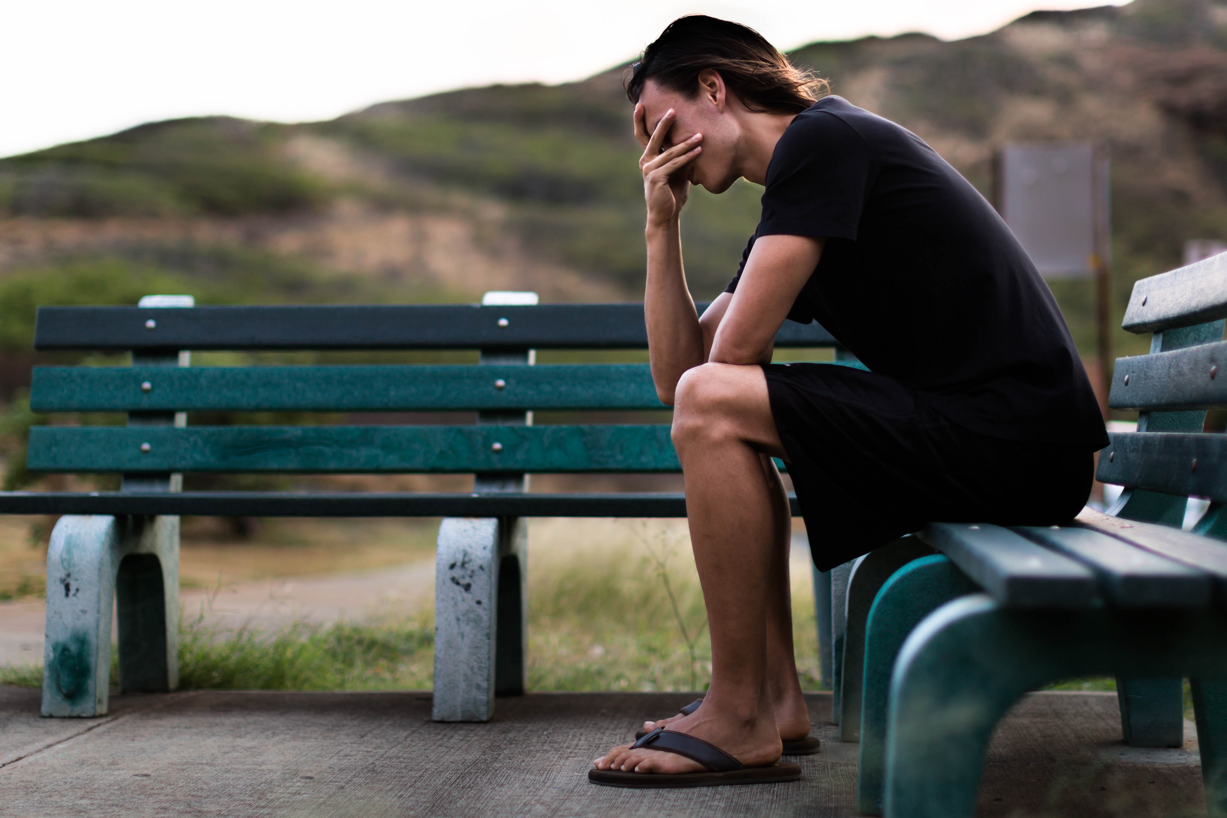 The image captures a young man sitting on a park bench, his head resting on his hand in a contemplative and distressed posture. He is dressed in casual black clothing and sandals, with his gaze directed downwards, suggesting a moment of introspection or emotional struggle. The background features a blurred landscape of hills and greenery, indicating a natural setting, while the benches provide a public park atmosphere.