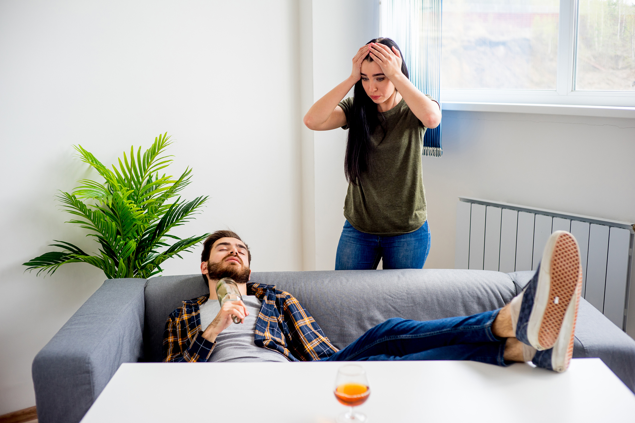 The image depicts a couple in a domestic setting. A man is reclining on a grey sofa, appearing relaxed or possibly intoxicated, with a bottle in his hand. A woman stands nearby, visibly distressed, with her hands on her head, indicating concern or frustration. The room is bright and features a large window, a green plant, and simple furnishings.