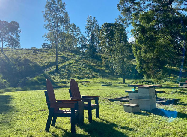 The image depicts a serene outdoor setting that perfectly embodies the charm of Yalbaroo Scenic Rim Retreat. Two rustic wooden chairs are positioned invitingly on a lush green lawn, providing a perfect spot for guests to relax and soak up the tranquil surroundings. The sunlight filters through the trees, casting a warm glow over the landscape, which features rolling hills in the background and natural greenery typical of the Scenic Rim area. In the foreground, a stone fire pit is visible, suggesting an ideal place for evening gatherings or marshmallow toasting under the stars. This image evokes a sense of peace and connection with nature, highlighting the retreat's focus on providing a serene holiday accommodation experience for visitors seeking relaxation and outdoor adventure in the beautiful Cainbable region and surrounding areas.