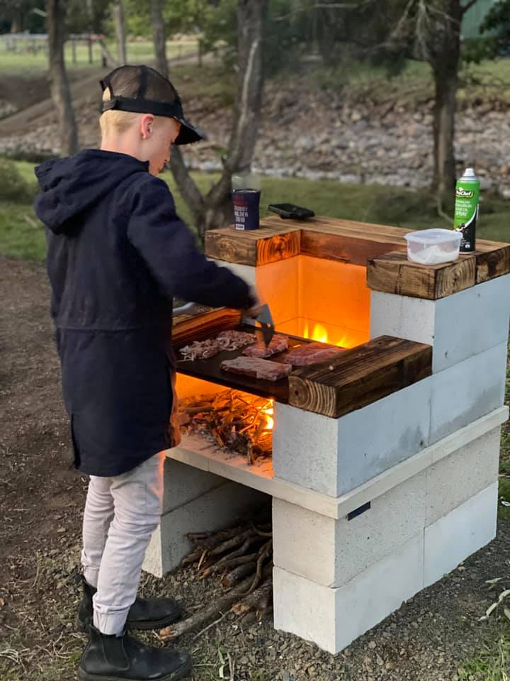 In this inviting scene at the Yalbaroo Scenic Rim Retreat, a young child is focused on cooking at a rustic outdoor barbecue station. The grill is made from a combination of wooden and concrete materials, providing a sturdy cooking surface where several pieces of meat sizzle over glowing embers, suggesting a cozy night of outdoor cooking. The backdrop features a serene natural setting, with trees and a riverbed visible in the background, enhancing the retreat's tranquil atmosphere. This setting is perfect for families wishing to bond over a barbecue in the beautiful surroundings of Cainbable, making it an ideal choice for holidaymakers looking for a relaxing getaway in the Scenic Rim.