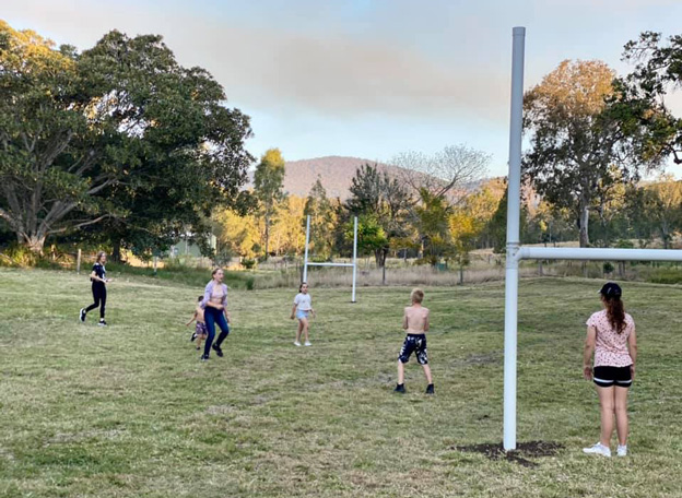 The image shows a lively outdoor scene at Yalbaroo Scenic Rim Retreat, where a group of children are engaged in a game of football on a grassy field surrounded by lush trees. The goalposts, set up for the game, add to the recreational ambiance. The landscape is picturesque, featuring the rolling hills and natural beauty typical of the Scenic Rim region, making it an ideal setting for families looking for holiday accommodation that offers both relaxation and outdoor activities. The children, dressed casually, appear energetic and joyful, reflecting a fun and active atmosphere that holidaymakers at Yalbaroo would undoubtedly enjoy. The retreat likely provides access to such recreational opportunities, catering to families and groups seeking an escape into nature.